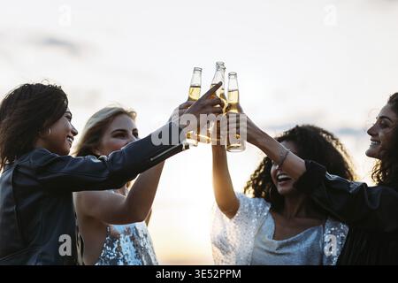 Bei Sonnenuntergang stoßen Freunde aus verschiedenen Ethnien Bier an. Frauen feiern abends mit Bier Stockfoto