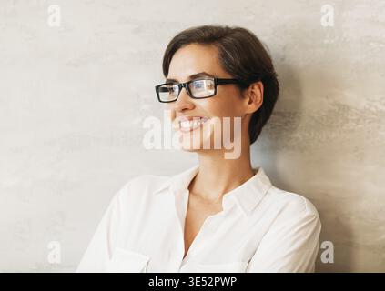 Fröhliche Geschäftsfrau mit Brille. Porträt eines lächelnden Unternehmers, der sich in formeller Kleidung an eine Wand lehnt Stockfoto