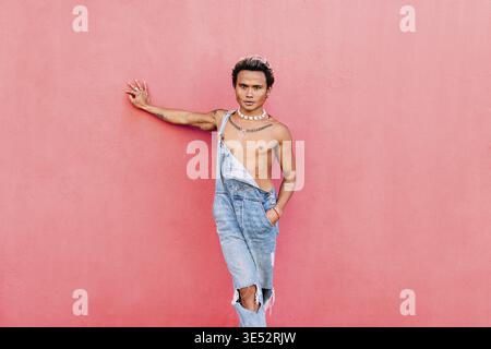 Ein junger, hübscher Mann, der Jeans trägt und an der rosa Wand angelehnt ist und die Kamera anschaut Stockfoto