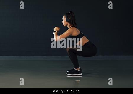 Seitenansicht der muskulösen Frau mit Kopfhörern tun hockt in Fitnessraum Stockfoto