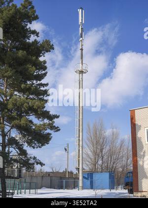 Ein hoher Mobilfunkturm mit Antennen erhebt sich vor einem hellblauen Himmel, der von Wolken gesäumt ist, in einer Winterlandschaft Stockfoto