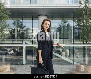 Ernsthafte und selbstbewusste Frau mittleren Alters in formeller schwarzer Kleidung, die vor einem modernen Geschäftsgebäude steht Stockfoto