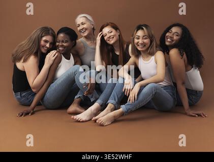Sechs Frauen unterschiedlichen Alters sitzen zusammen im Studio auf braunem Hintergrund. Multiethnische Gruppe von Frauen mit guten Zeiten Stockfoto