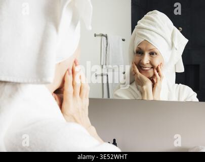 Fröhliche Frau bewundert ihr Spiegelbild im Badezimmerspiegel. Reife Frau, die ihr Gesicht nach dem Heim-Spa mit den Händen berührt Stockfoto