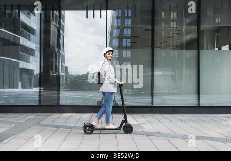 Seitenansicht einer jungen lächelnden Geschäftsfrau mit Fahrradhelm auf dem Kopf, die einen elektrischen Motorroller fährt Stockfoto