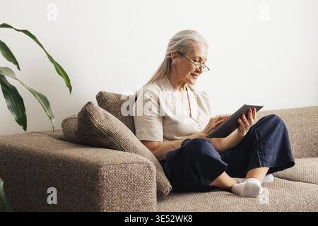 Alte Frau, die mit einem digitalen Tablet auf einer Couch sitzt Stockfoto