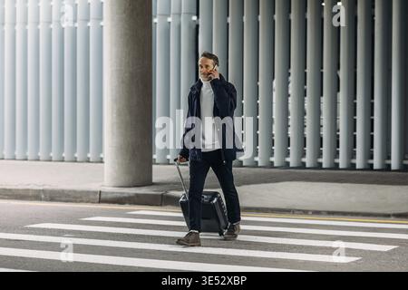 Ein älterer Mann spricht auf dem Handy, während er auf einem Fußgängerüberweg spaziert und Koffer zieht Stockfoto