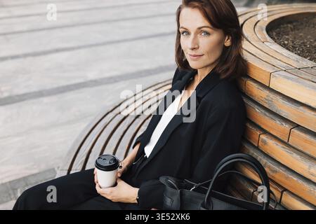 Lächelnde Geschäftsfrau mittleren Alters, die draußen auf einer Bank sitzt. Frau in Schwarz formell tragen entspannende Outdoor-Kleidung mit einer Tasse Kaffee Stockfoto