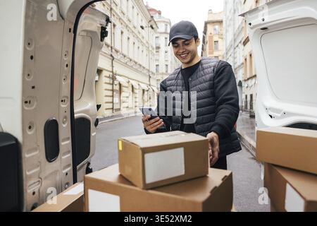 Lächelnder Kurier in Uniform, der am Kofferraum des Autos stand und das Mobiltelefon auf Kartons blickte Stockfoto