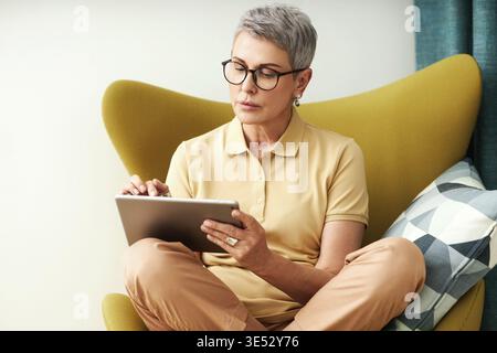 Reife Frau, die mit überkreuzten Beinen auf einem Sessel sitzt, mit einem digitalen Tablet Stockfoto
