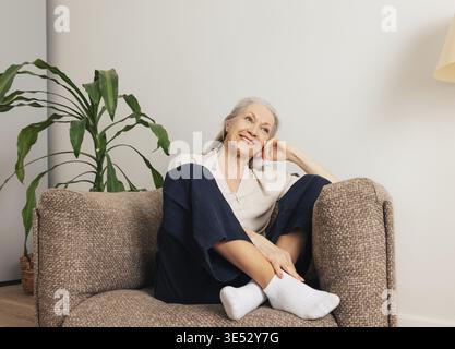 Fröhliche Seniorin, die sich zu Hause in einem Sessel entspannt. Lächelnde alte Frau in den Casuals im Wohnzimmer Stockfoto