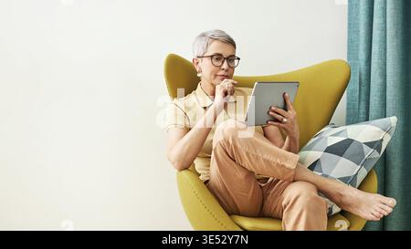 Stilvolle, reife Frau mit Brille, die sich zu Hause ein digitales Tablet anschaut Stockfoto