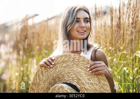 Junge schöne Frau mit einem großen Strohhut auf einem Feld, umgeben von hohem Gras Stockfoto