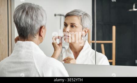 Schöne Seniorin im Bademantel, die vor einem Spiegel steht Stockfoto