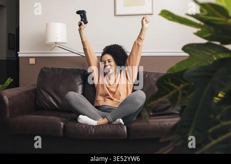 Glückliche Mixed-Race-Frau, die auf einem Sofa im Wohnzimmer sitzt, mit erhobenen Händen Stockfoto