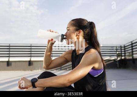 Seitenansicht einer jungen Frau in Sportbekleidung, die Trinkwasser aus einer weißen Flasche auf dem Dach trägt Stockfoto