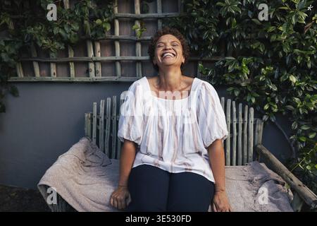 Frau mittleren Alters lacht, während sie draußen auf einer Bank im Garten sitzt Stockfoto