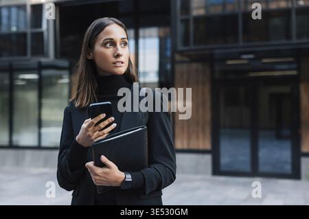 Selbstbewusste und seriöse Frau in schwarzer Kleidung, die ein Smartphone und ein digitales Tablet vor einem Glasgebäude hält. Junge Geschäftsfrau trägt Stockfoto