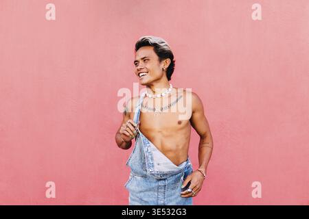 Porträt eines jungen lächelnden Mannes in Denim-Overalls, der wegschaut, während er an einer rosa Wand steht Stockfoto