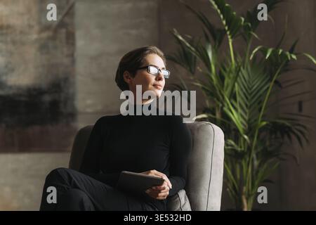Junge Frau in schwarzer formeller Kleidung trägt eine Brille und hält ein digitales Tablet, während sie in ihrem Loft sitzt Stockfoto