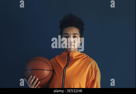 Porträt eines jungen Basketballspielers in oranger Sportkleidung, der im Studio posiert. Frau mit einem Basketball, der vor blauem Hintergrund steht Stockfoto