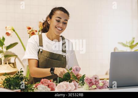 Lächelnde Frau, die in ihrem Blumenladen arbeitet. Blumenläuferin, die Blumen auf der Theke arrangiert Stockfoto