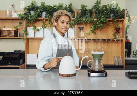 Porträt eines jungen Barista an einem Tresen in einem Café. Junge Frau, die als Barista arbeitet und einen Wasserkocher hält und in die Kamera schaut Stockfoto
