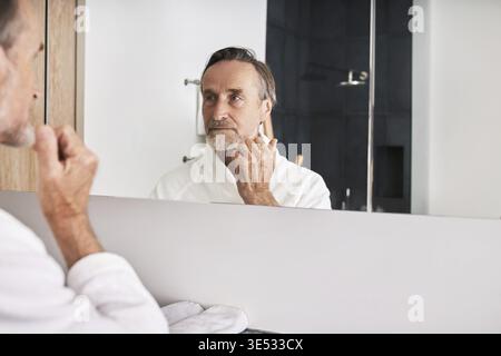 Gutaussehender älterer Mann mit weißem Bademantel untersucht seine Haut morgens im Badezimmer Stockfoto