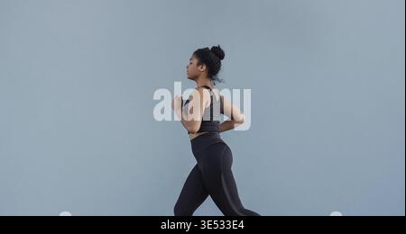 Seitenansicht einer Frau in schwarzer Sportswear, die an der grauen Wand läuft. Junge Frau, die draußen joggen Stockfoto