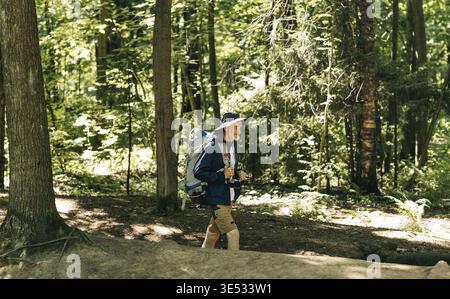 Seitenansicht eines älteren Mannes mit Rucksack und Wanderkleidung, der an sonnigem Tag durch den Wald spaziert Stockfoto