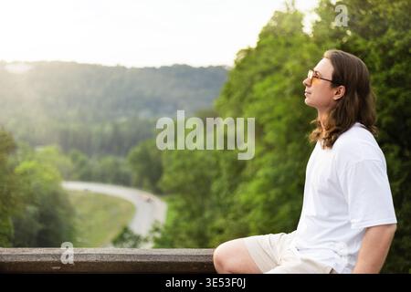 Profil eines entspannten jungen Mannes mit langen Haaren und Sonnenbrille, der auf einer Steinmauer sitzt und einen malerischen Blick auf eine gewundene Straße und den üppigen grünen Wald A genießt Stockfoto