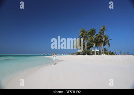 Blonde Frau im türkisfarbenen Kleid geht barfuß entlang der weißen Sandbar neben der Calm Turquoise Lagoon in Richtung luxuriöser Overwater Villas auf den Malediven Stockfoto