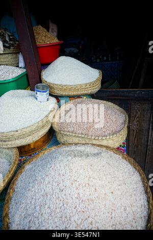 Körbe gefüllt mit verschiedenen Reissorten an einem Marktstand, mit etwas getrocknetem Fisch im Hintergrund. Stockfoto