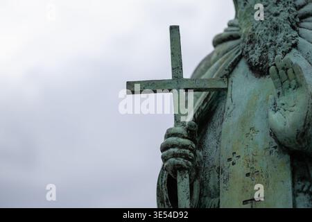 Nahaufnahme einer verwitterten Bronzestatue, die ein einfaches Kreuz gegen einen stimmungsvollen Himmel hält, mit komplizierten Grüngris-Texturen und einer symbolischen religiösen Geste Stockfoto