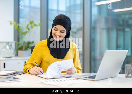 Muslimische Geschäftsfrau im Hijab arbeitet an ihrem Schreibtisch in einem modernen Büro, liest Papiere und unterzeichnet gerne ein Dokument mit einem Stift, Laptop und Karten sichtbar Stockfoto