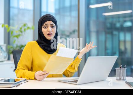 Muslimische Geschäftsfrau mit Hijab, die an ihrem Schreibtisch sitzt und sich verwirrt und frustriert fühlt, während sie einen Umschlag mit offiziellen Dokumenten hält und auf unerwartete schlechte Nachrichten oder ein Problem reagiert Stockfoto