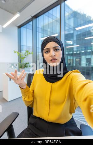 Muslimische Geschäftsfrau mit Hijab und gelbem Hemd, die in einem modernen Büro sitzt, ihr Smartphone für einen wichtigen Videoanruf benutzt und während der Kommunikation Verwirrung ausdrückt Stockfoto