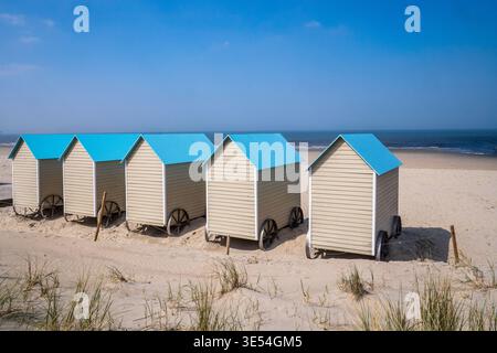 Fünf beigefarbene hölzerne Strandhütten mit hellblauen Dächern, die auf großen schwarzen Rädern montiert sind. Die Gebäude liegen auf dem goldenen Sand, der auf das ruhige Meer blickt Stockfoto
