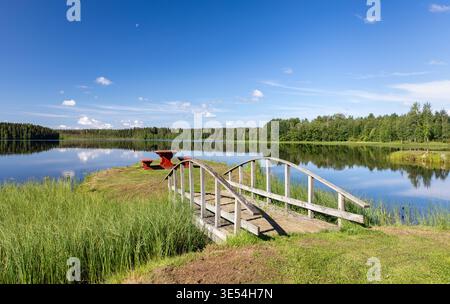 Wunderschöne und friedliche sommerliche Seeszene an einem sonnigen Morgen in Finnland. Die Brücke führt über das Wasser zu einer kleinen Insel, wo sich ein Platz befindet. Weitwinkel. Stockfoto