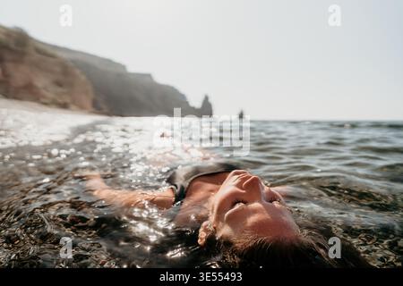 Frau schwimmendes Wasser Frau entspannt sich im flachen kristallklaren Küstenmeer mit schimmerndem Sonnenlicht und Klippen Stockfoto