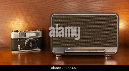 Ein kabelloser Bluetooth-Lautsprecher im Retro-Stil und eine Vintage-Kamera mit warmer Beleuchtung auf einem Holzregal Stockfoto
