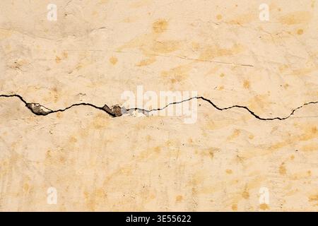 Gerissene beige Putzwand mit dunkler Spalte, robuste verwitterte Oberfläche mit gesprenkelten Flecken und kleinen Ablagerungen, starker horizontaler Rissschnitt Stockfoto