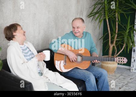 Ältere Ehepaare verbringen Zeit miteinander. Gesunder Lebensstil. Emotionale Gesundheit. Rest von Gadgets. Ein älterer Mann spielt Gitarre für seine Frau, die zu Hause auf der Couch sitzt. Alles Gute zum Valentinstag || Model veröffentlicht Stockfoto