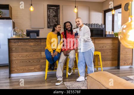 Verschiedene Freunde, die sich mit dem Smartphone an der Holztheke auf gelben Hockern unterhalten und Senftop tragen. Café, Kaffee, Gruppe, Telefon, Konversation, Ambiente, modern || Modell veröffentlicht Stockfoto