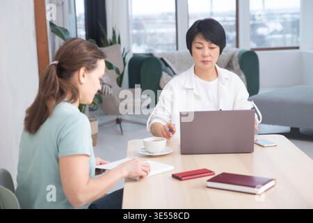 Glückliche, multiethnische Kollegen sitzen zusammen am Coworking-Platz, erfahrenere Mitarbeiter teilen sich. Die entzückende asiatische Geschäftsfrau berät den Kunden im Büro || Model veröffentlicht Stockfoto