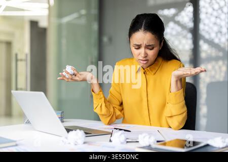 Gestresste Geschäftsfrau, die mit einem Büroproblem zu tun hat, zerknittertes Papier hält und Frustration zeigt, während sie mit einem Laptop und Dokumenten an ihrem Schreibtisch sitzt Stockfoto