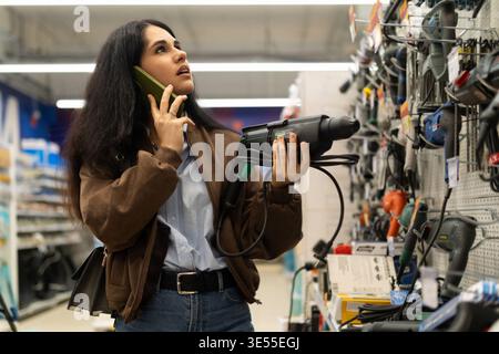Eine Frau, die einen Bohrer hält und in einem Eisenwarenladen auf ihrem Smartphone spricht Stockfoto