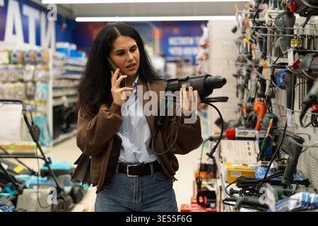 Frau im Eisenwarengeschäft, die einen Bohrschrauber hält, während sie mit dem Smartphone spricht Stockfoto