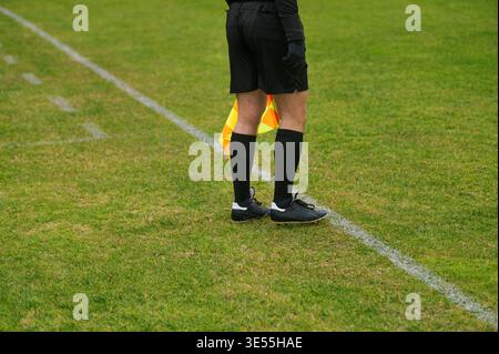 Der Fußballschiedsrichter steht auf der Grasfeldlinie. Er hält die orangefarbene Flagge in der Hand. Schwarze Shorts und Socken runden seine Uniform ab. Die Schuhe liegen fest auf dem grünen tur Stockfoto