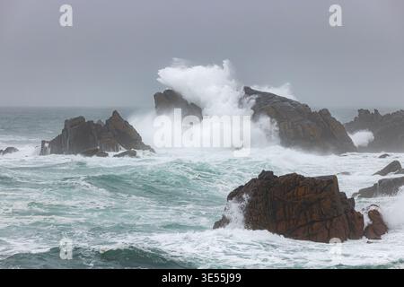Blick auf große Wellen, die gegen dunkle Küstenfelsen unter einem stürmischen, grauen Himmel in Ushant, Bretagne, Frankreich prallen. Stockfoto